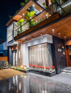 Modern two-story house with balcony, illuminated garden plants, and reflective driveway at night.